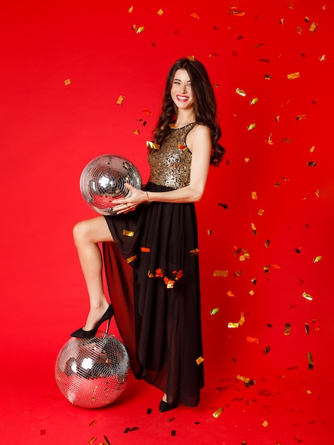 A brunette girl is standing in a black dress with gold sequins holding a disco ball in her hands