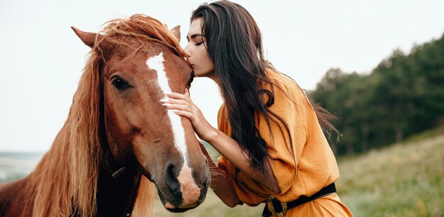 brunette girl kissing her horse while posing in a field