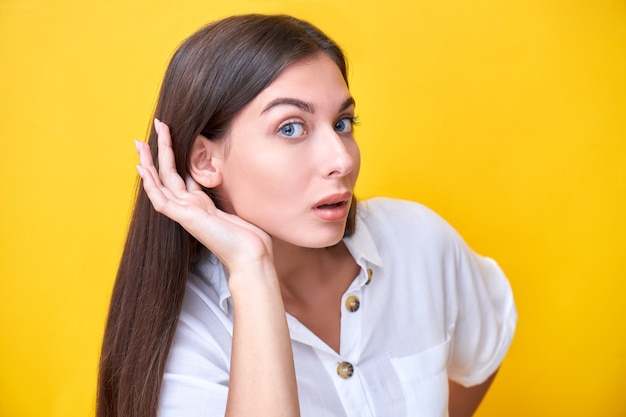 Brunette girl looks curiously at the camera with hand behind her ear, overhears something interesting isolated on yellow background