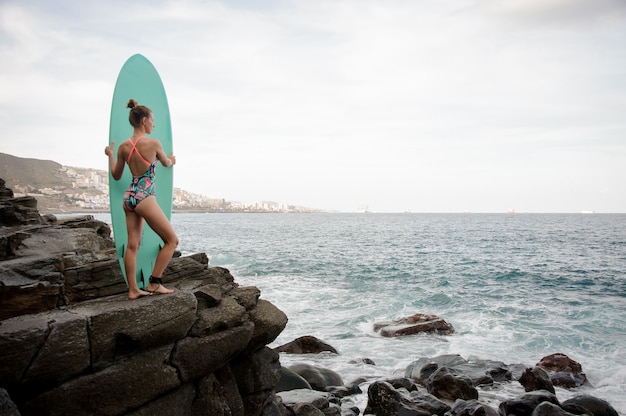 Brunette girl in the multi colored swimsuit standing with the surf on the rock of Atlantic ocean looking at the distance