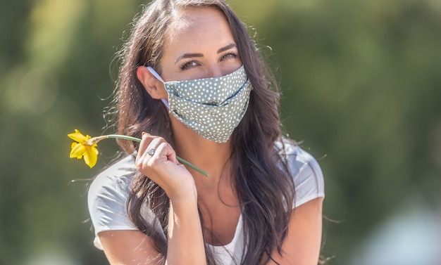 Brunette girl in the nature wears facemask smiling, holding a daffodil in her hand.