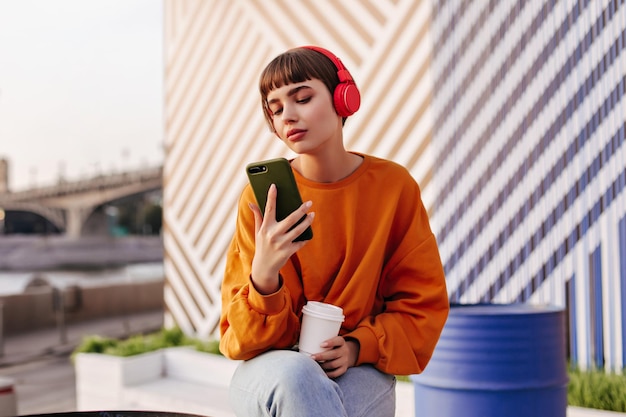 Brunette girl in orange sweatshirt holding phone outside Stylish woman in headphones listening to music on striped backdrop