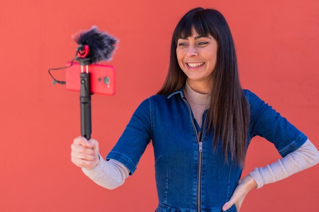 Brunette girl recording a vlog in a blue denim outfit. Caucasian pretty a red background