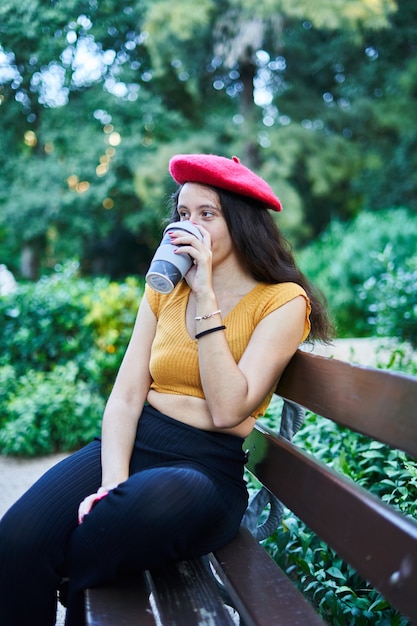 Ragazza castana in berretto rosso che si siede sulla panchina del parco a bere caffè