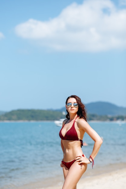 brunette girl in a red bikini swimsuit and sunglasses posing on the beach against the sea.