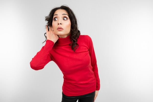 Brunette girl in a red sweater overhears a conversation on a white studio background.