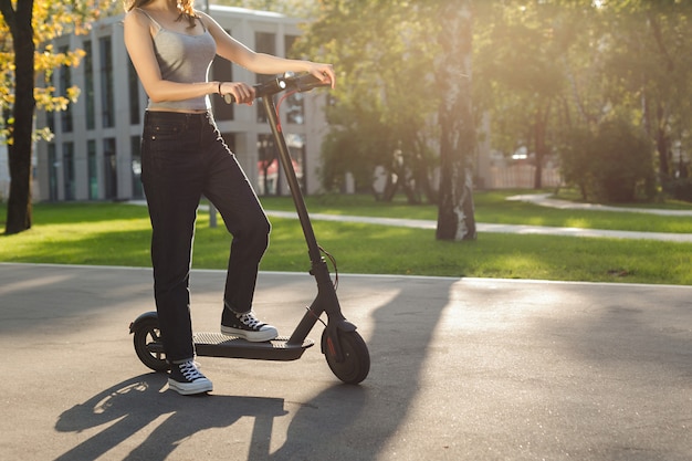 Photo brunette girl riding an ecofriendly electric kick scooter in a park in sunny weather on sidewalks