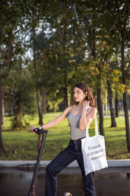 Brunette girl riding a scooter with a bag on his shoulder in the park in sunny weather with a smile