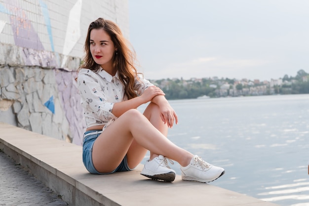 A brunette girl in shorts and sneakers walks by a large lake. Girl in short denim shorts on the waterfront of a city lake.