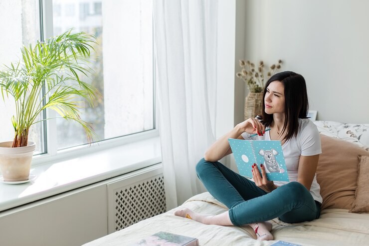 Девушка брюнетка сидит на кровати в светлой комнате, читая книгу.