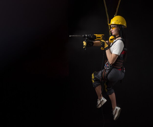 Брюнетка в спецодежде и рабочий в каске позирует на черном фоне с рабочим инструментом (девушка, висящая на альпинистском снаряжении, строительные работы на высоте)