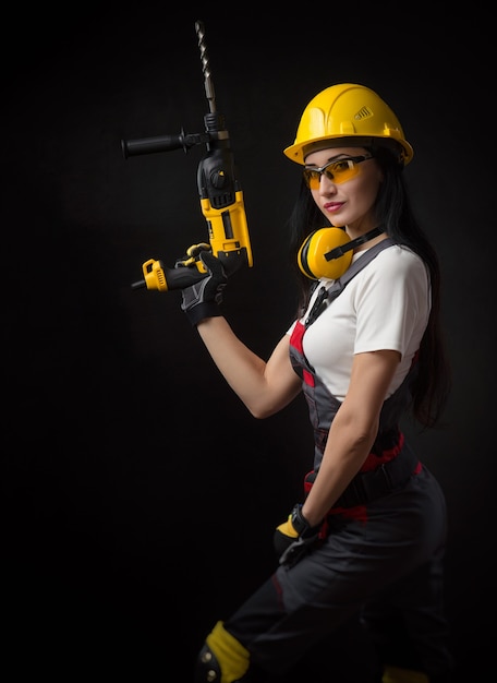 The brunette girl in special clothes and a worker in a helmet posing on a black background with a working tool ( perforator, drill, percussion hammer )