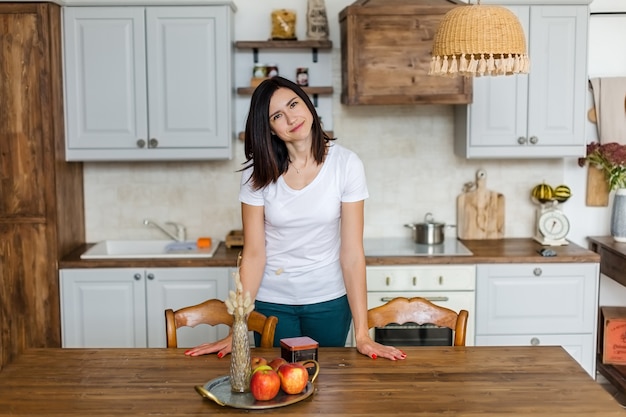 Brunette girl stands at the table in the kitchen looks into the frame. 