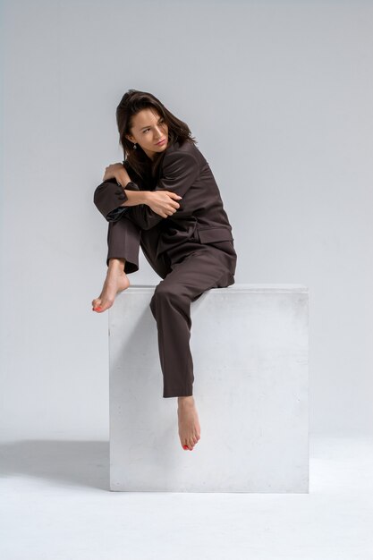 A brunette girl in a strict brown suit is sitting on a white cube on a white background.emotions of a businesswoman.