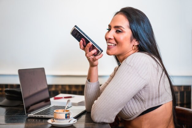 Ragazza bruna che telelavora in una caffetteria con il suo laptop, inviando un messaggio audio a un collega