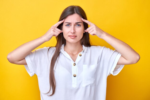 Brunette girl tensely holds fingers at temples thinks hard and makes a choice isolated on yellow background