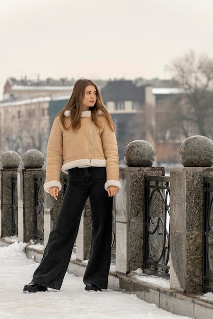 Brunette girl in trendy black jeans and beige sheepskin coat stands pensively on bridge Winter look for every day