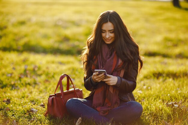 brunette girl using her phone as she is relaxing at the park during autumn