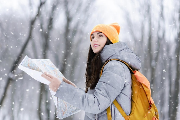 회색 재킷과 노란색 모자를 쓴 갈색 머리 소녀는 지도를 들고 겨울 눈 덮인 숲에서 배낭을 메고 걷는 동안 lanscapes를 고려합니다.