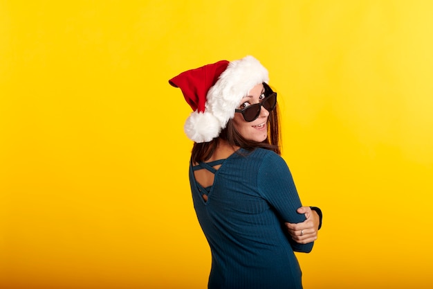 A brunette girl wearing a santa claus hat with a yellow background and sunglasses