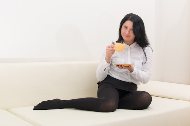 brunette girl in a white blouse sits on a sofa with a cup of coffee