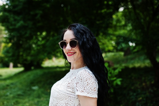 Brunette girl in white blouse with sunglasses posed at park.