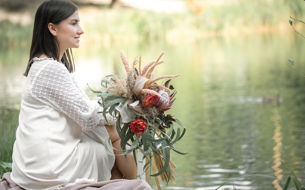 Una ragazza bruna in abito bianco si siede in riva al fiume con un mazzo di fiori esotici, sfondo sfocato.