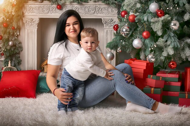 a brunette girl in a white Tshirt hugs her little son sitting by the Christmas tree fireplace