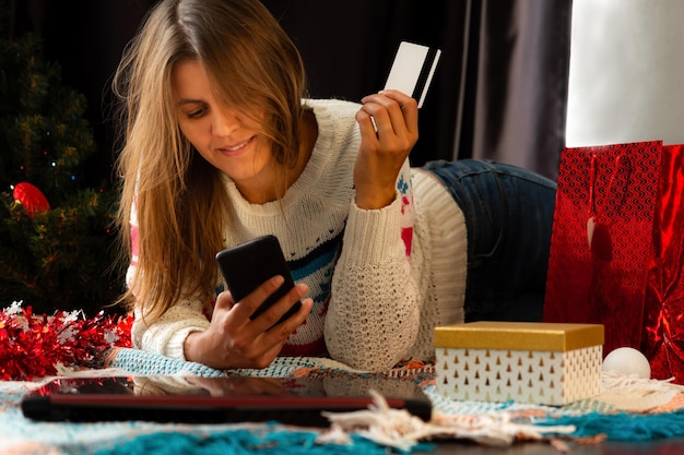 自宅で冬のセーターを着たブルネットの女の子が笑顔で携帯電話を見てクリスマスの買い物をする