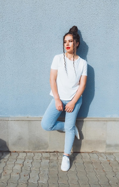 A brunette girl with dreadlocks and red lipstick in a denim suit is standing near a blue wall generation z