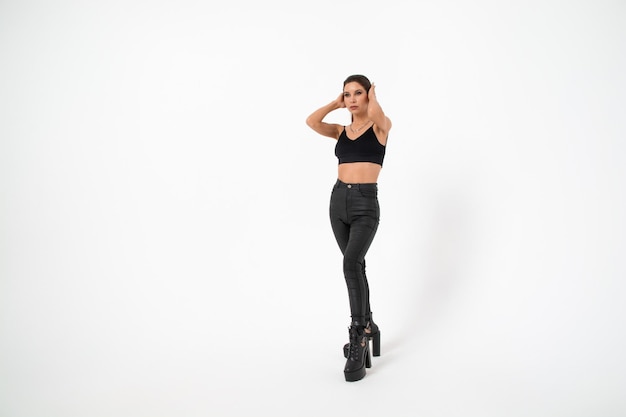 A brunette girl with long hair in black leather pants and boots poses on a white backgroundyouth fashion