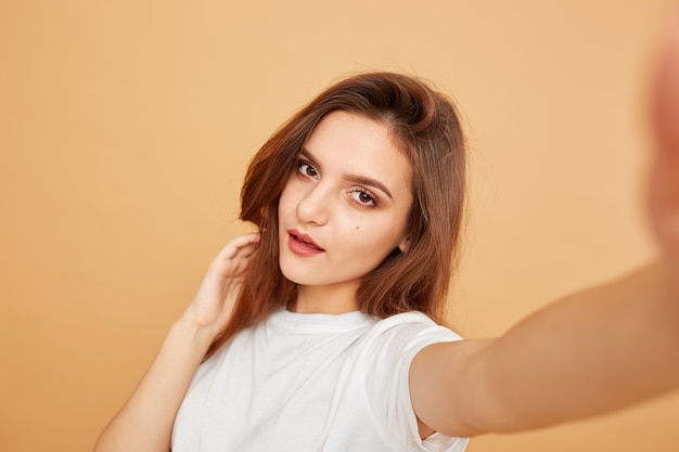 Brunette girl with long hair dressed in white t-shirt makes a selfie on the beige background in the studio .