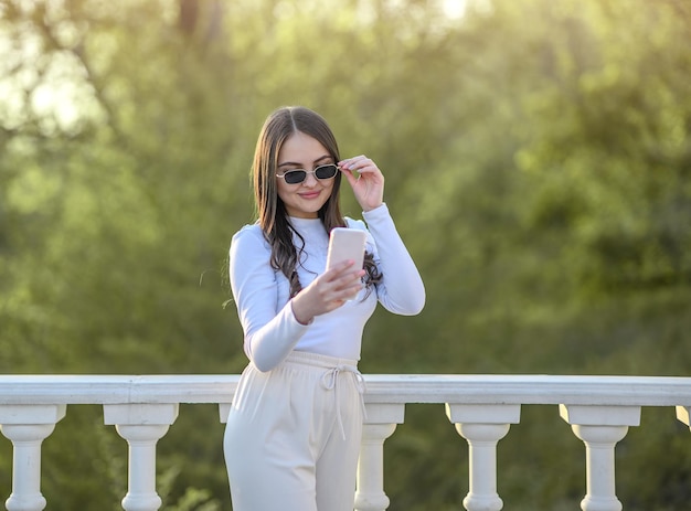 Ragazza bruna con i capelli lunghi con gli occhiali e con un telefono si fa un selfie per strada