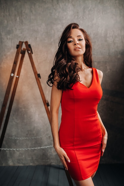 A brunette girl with long hair in a red dress poses in the studio.