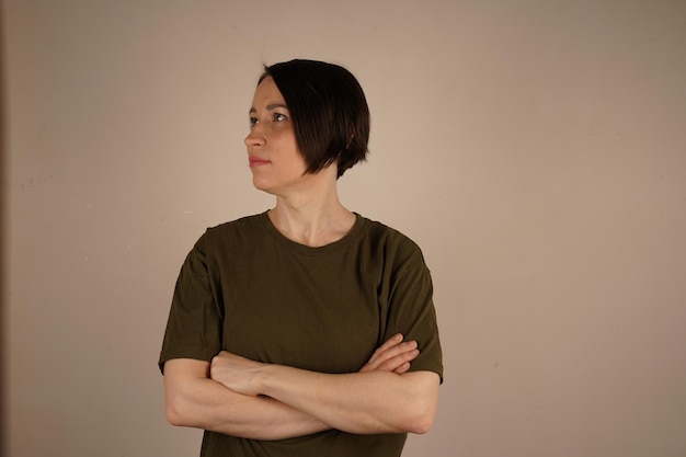 A brunette girl with short hair and gray eyes She crossed her arms in front of her and looks in the other direction The girl is found On a beige background