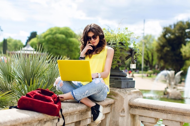 Brunette girl working with yellow laptop on summer in park. She is sitting on fence, looking busy.