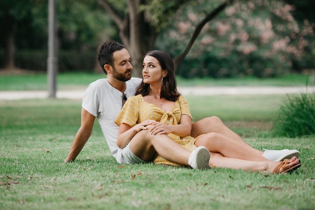 A brunette girl in a yellow dress is sitting between the legs of her boyfriend on the grass in old Spain town. A couple of tourists on a date in the Valencian park.