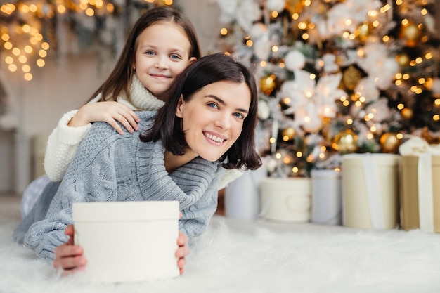 Brunette glimlachende vrouwelijke volwassene en haar mooie schattige kleine kind in witte trui in de woonkamer houden aanwezig om vader en echtgenoot te feliciteren met komend nieuwjaar of kerstmis