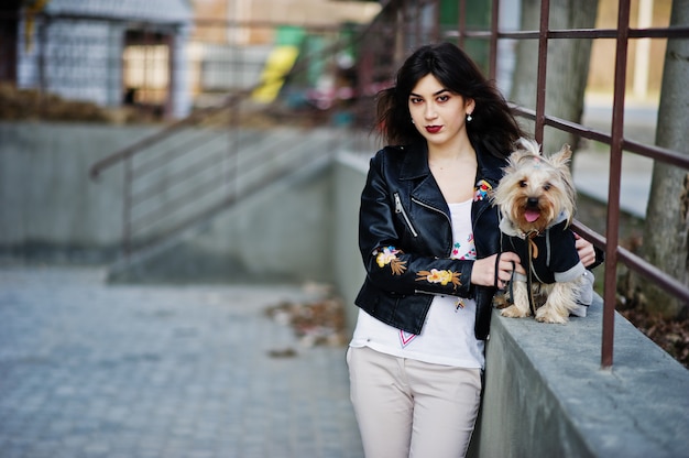 Brunette gypsy girl with yorkshire terrier dog posed against steel railings. Model wear on leather jacket with ornament, pants.