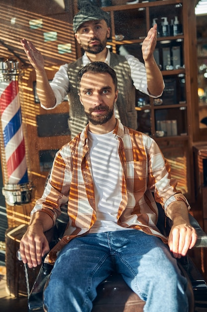 Brunette hair-stylist holding hands up while his client is sitting in an armchair at a barbershop