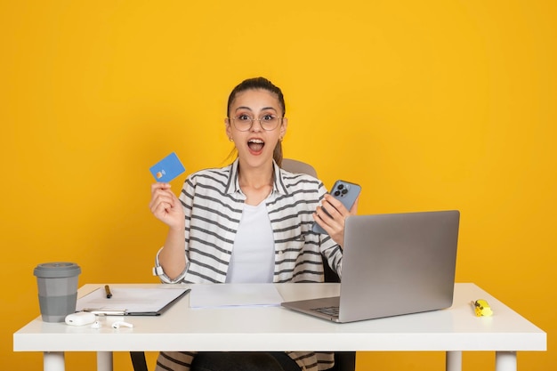 Brunette happy business woman sit at office desk using laptop holding credit card and phone