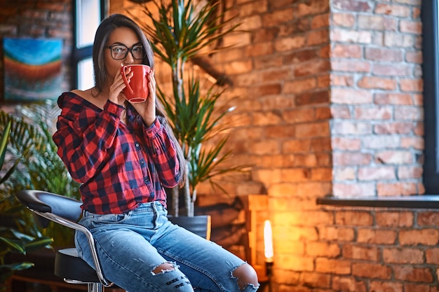 A brunette hipster female dressed in a fleece shirt and jeans drinks coffee in a room with loft interior.