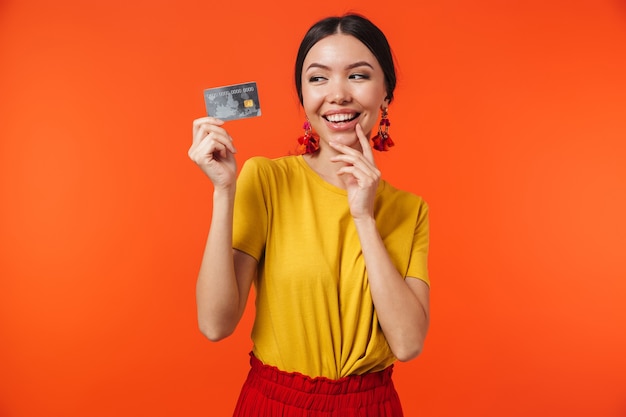 Foto bruna donna ispanica 20s vestita con gonna sorridente e tenendo la carta di credito isolate su muro rosso