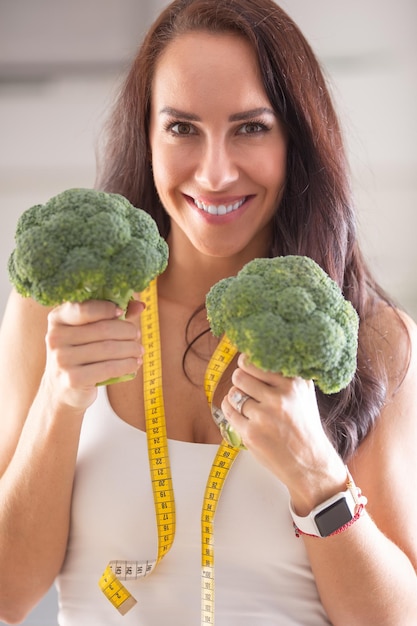 Brunette holding two brocollis as a symbol of healthy lifestyle with measuring tape around her neck