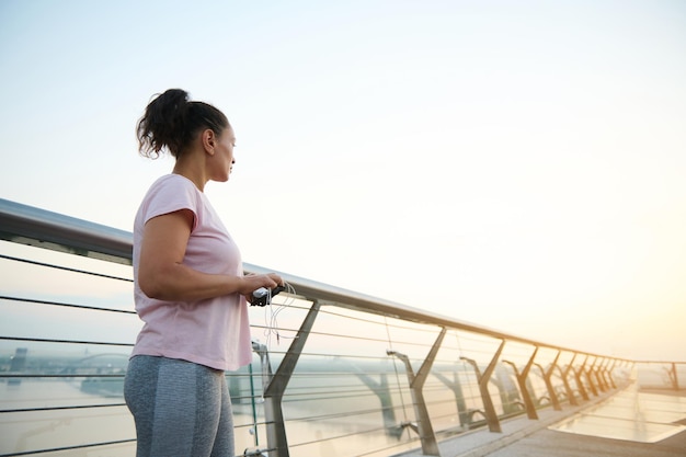 Brunette in roze T-shirt en grijze sportlegging houdt koptelefoon in haar handen en kijkt in de verte, staande op de stadsbrug, klaar voor een ochtendrun. Prachtige zonsopgang op een loopband