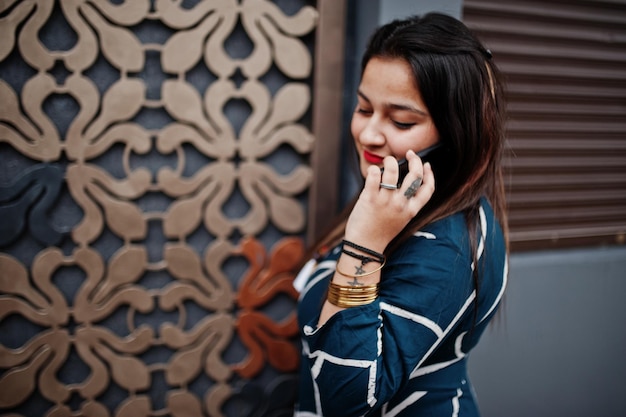 長いファッショナブルなドレスを着たブルネットのインドの女性が屋外でポーズをとり、携帯電話で話す