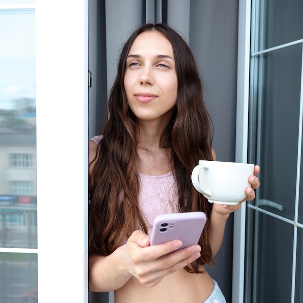The brunette is drinking coffee on the balcony and texting on the phone A relaxed weekend morning