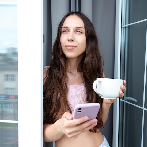 ブルネットはバルコニーでコーヒーを飲み、電話でテキストメッセージを送っていますリラックスした週末の朝