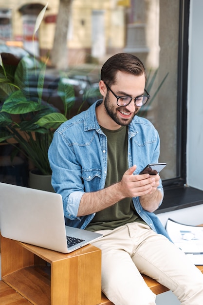 brunette jonge man met een bril die een smartphone vasthoudt terwijl hij in de buitenlucht op een laptop in het stadscafé werkt