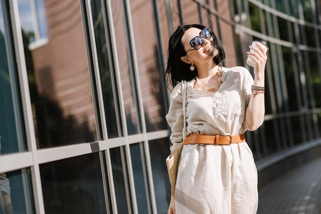 Brunette jonge vrouw met zonnebril en tas met koffie wandelen in de stad. Lifestyle portret van vrouw